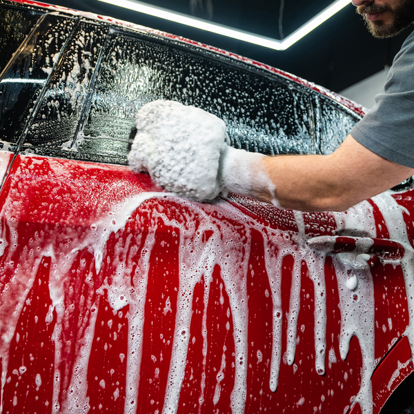 Car detailing: Red auto is being washed with soap and mitt by a man, under bright lights. Car care.